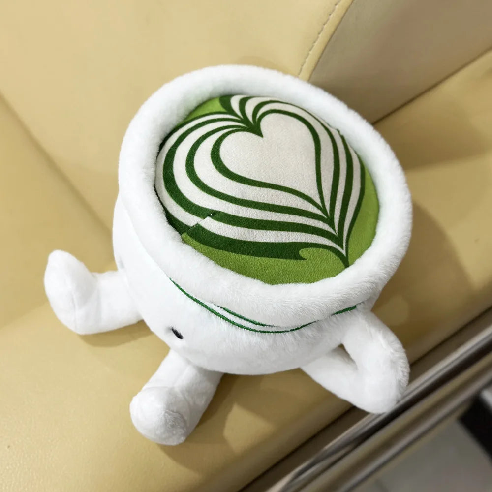 Cute matcha plushie sitting on wooden desk next to real matcha latte
